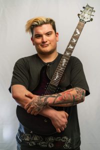 Bailey Chamberlain with his arms wrapped around his guitar placed in front of him.