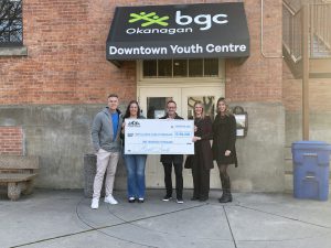 This photograph shows the two entrepreneurs holding a giant cheque with some of the BGC Okanagan leadership team. The giant cheque represents the donation of $100,000. The group is standing in front of the doors of the Downtown Youth Centre.