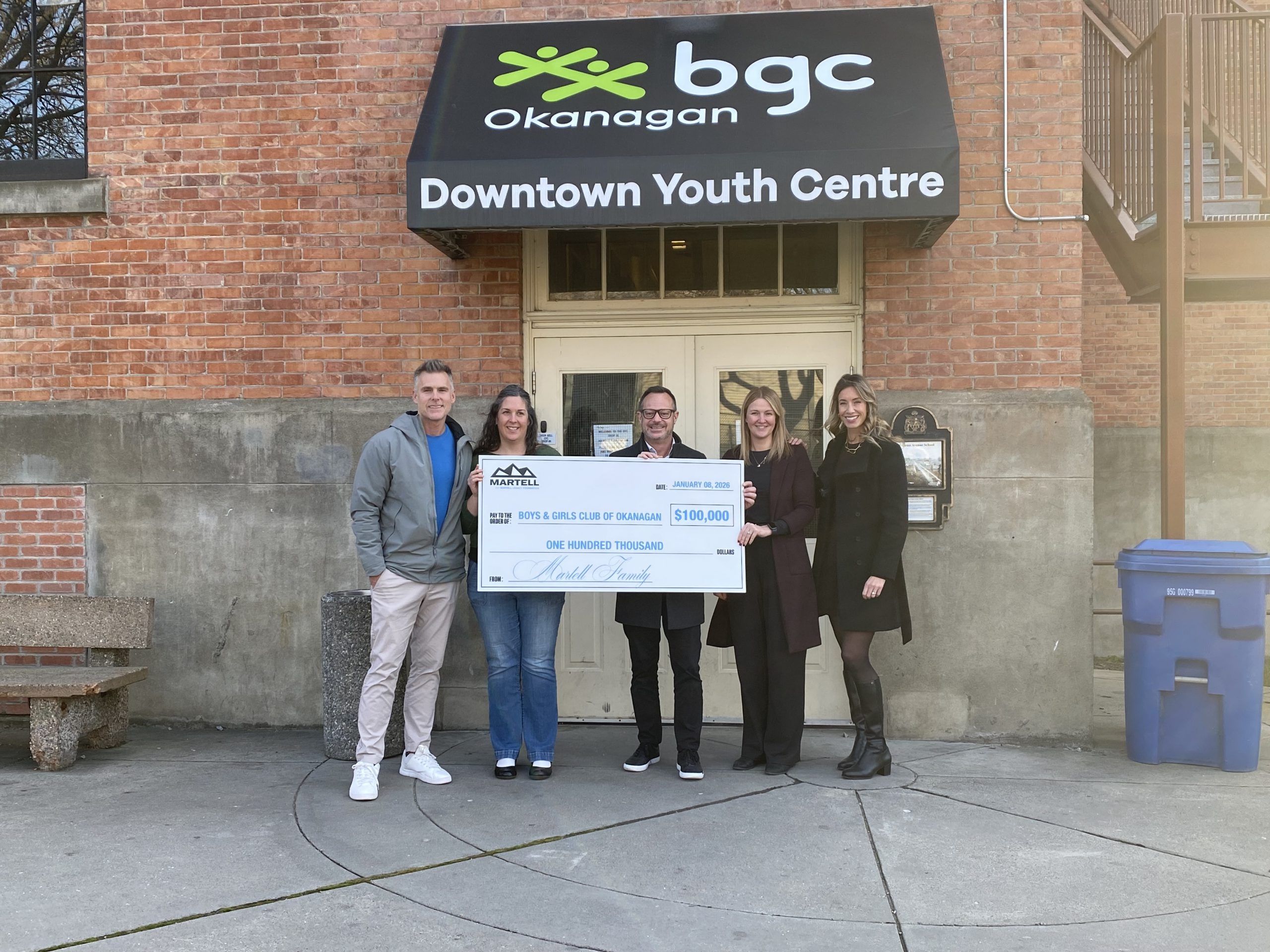 This photograph shows the two entrepreneurs holding a giant cheque with some of the BGC Okanagan leadership team. The giant cheque represents the donation of $100,000. The group is standing in front of the doors of the Downtown Youth Centre.